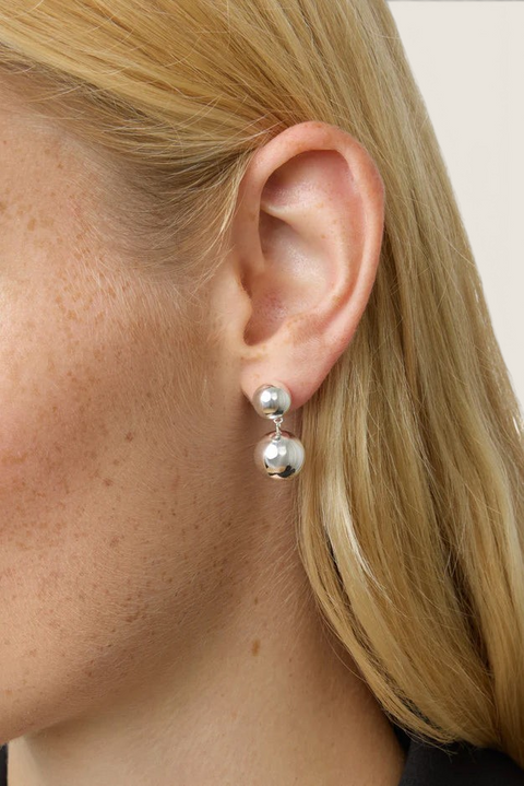 Bolina The Caroline silver earrings featuring a polished round stud and a dangling spherical charm on a woman's ear.